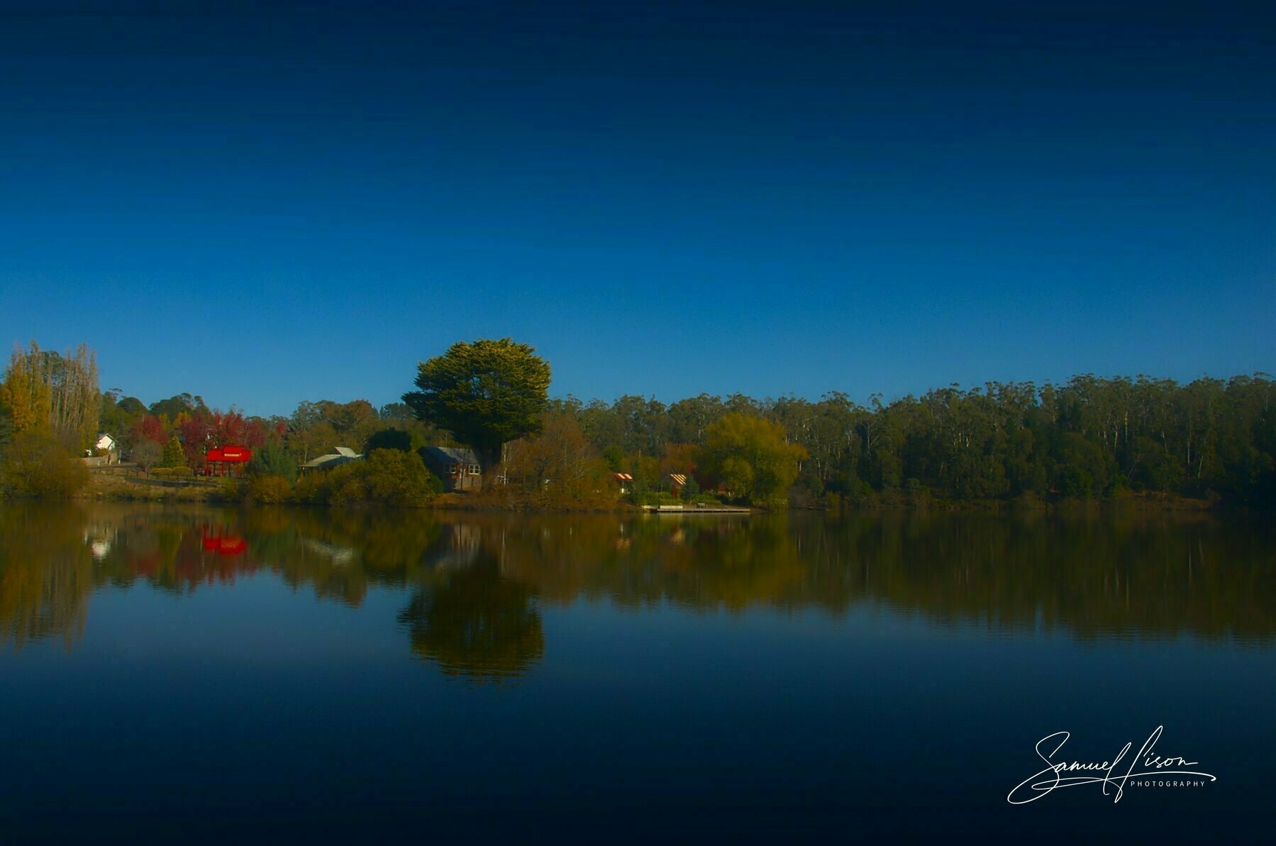 Auto-generated description: A serene lake reflects a tree-lined shore and vibrant autumn foliage under a clear blue sky.