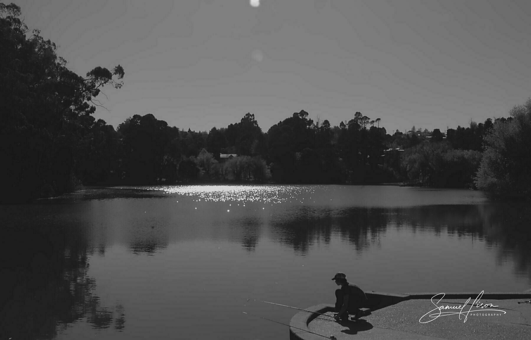 A person is fishing on the edge of a calm lake surrounded by trees, with the water reflecting sunlight.