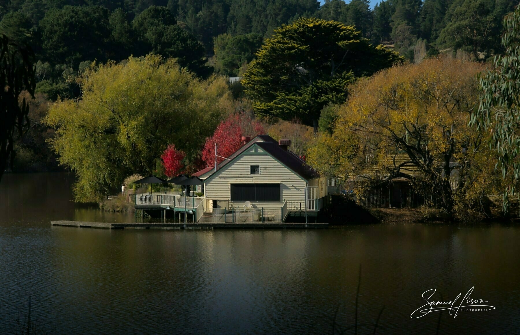 A boat shed sitting on Lake Daylesford, reflecting in the serene waters of the lake. Autumn trees of various colours surround the shed.