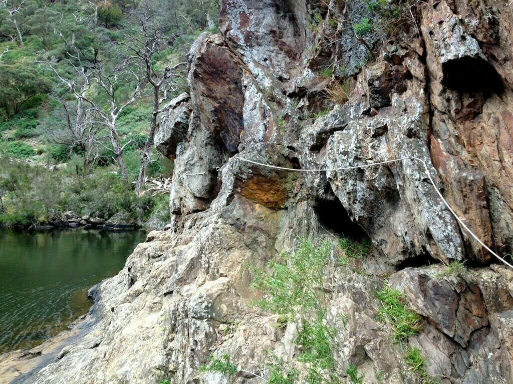 A rocky cliffside with a rope trail runs alongside a calm river with dry trees and greenery in the background.