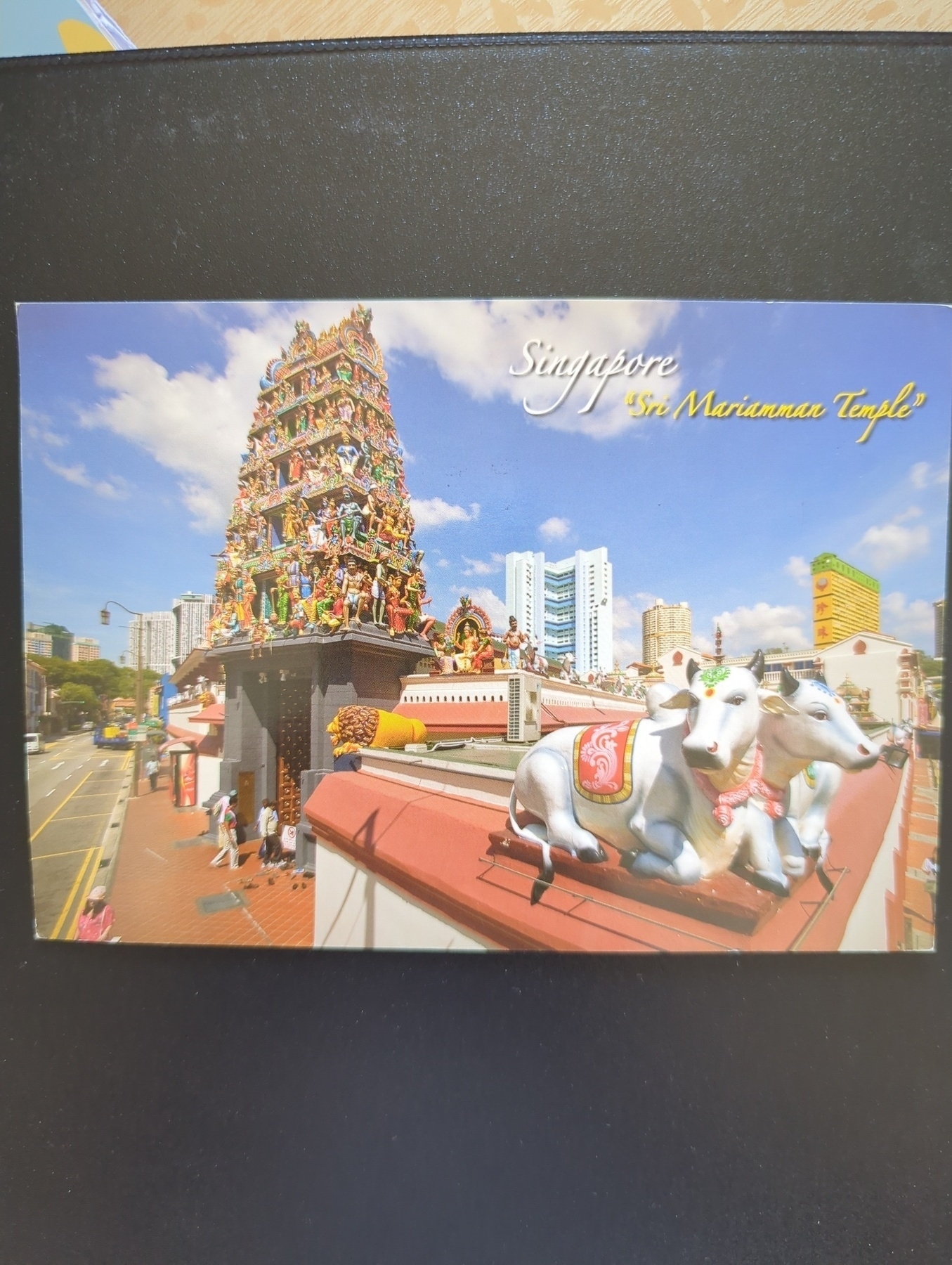 A vibrant postcard features the Sri Mariamman Temple in Singapore with intricate sculptures and two white statues of oxen in the foreground.