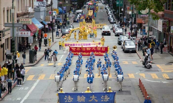 A marching band in blue uniforms leads a street parade with large banners and various performers, including people in yellow costumes. These are practitioners of the Falun Dafa faith, and they are marching through a city as part of a yearly World Falun Day celebrations.