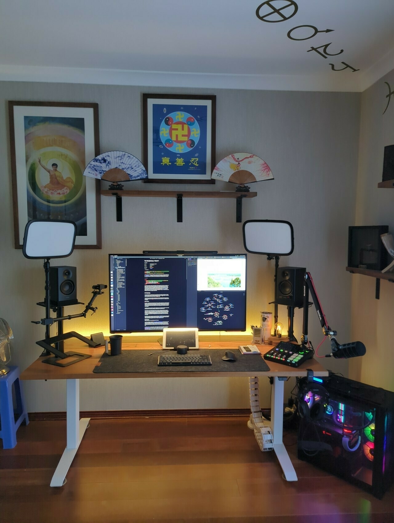 Photo of my Office Setup A tidy home office with a wooden desk, large monitor, tablet, keyboard, and mouse. Two LED panel lights, studio speakers, and a microphone are set up for recording. A colourful, illuminated PC is on the floor. Above the desk are three framed artworks and decorative fans on a shelf. Astronomy and astrological symbols are painted on the ceiling.