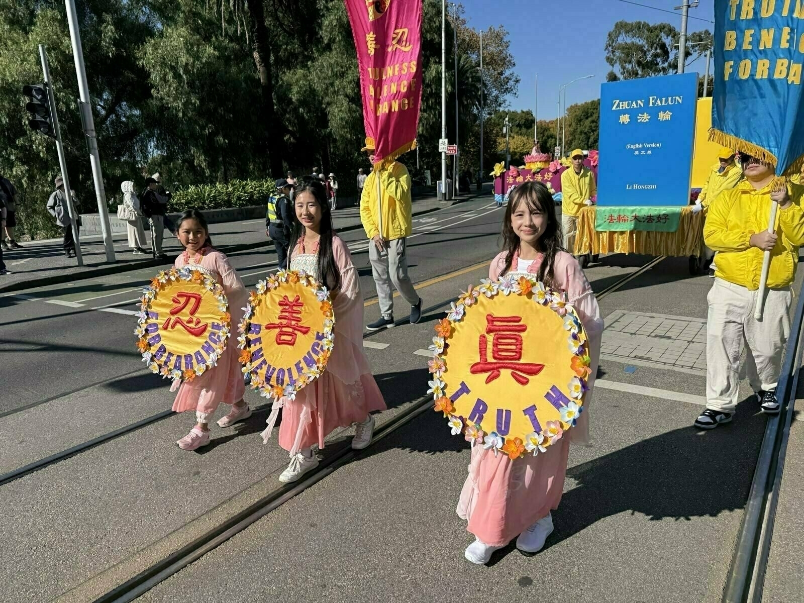 Three people in pink dresses hold round signs with the words TRUTH, COMPASSION, and TOLERANCE while participating in a parade.