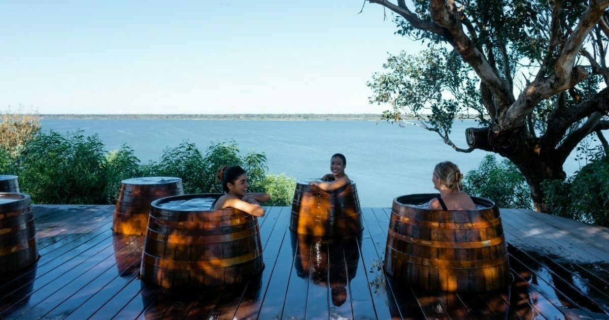 Metung Hot Springs Barrels Three people are relaxing in wooden hot tubs on a deck overlooking a body of water. These hot tubs are part of the Metung Hot springs and are naturally heated. The sulphur gasses cause random bubbles like a mini spa bath at times.
